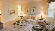 This single-floor residence exudes a warm and inviting ambiance, thoughtfully designed with comfort and elegance in mind. The living room serves as the heart of the space, showcasing soft, neutral wall tones that create a calming atmosphere. Plush wall-to-wall carpeting adds warmth underfoot, complementing the refined furnishings throughout. A generously sized sofa in a classic cream upholstery anchors the seating area, paired with a striking wrought iron and glass coffee table that introduces an artisan touch. Accent side tables with decorative table lamps cast a gentle, ambient glow, enhancing the room's welcoming feel. The walls are adorned with framed botanical and nature-inspired artwork, adding visual interest and a timeless aesthetic. A patterned accent chair provides an elegant contrast to the neutral palette. An open passageway connects seamlessly to adjacent areas of the home, contributing to an easy, flowing layout. The overall design balances classic furnishings with subtle decorative details, making this a comfortable and polished living space well-suited for both relaxing and entertaining.