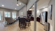 Located at 700 Martin Creel Blvd in Summerville, South Carolina, this single-story community space is defined by its dramatic vaulted ceilings and open-concept layout that seamlessly connects multiple functional zones. Light wood-look flooring runs throughout, complementing the neutral gray wall palette and creating a cohesive, contemporary aesthetic.

The lounge area near the entrance features large windows with plantation shutters that invite natural light and provide views of the surrounding greenery. Comfortable accent chairs arranged around a glass-topped coffee table create an inviting gathering space ideal for relaxed conversations or informal meetings.

Adjacent to the lounge, a high-top dining table with upholstered chairs serves as a central hub for group seating. The vaulted ceiling above this zone is accentuated by a pendant ceiling fan, enhancing the sense of openness and airflow throughout the space.

The kitchenette is well-appointed with rich dark espresso cabinetry, granite countertops, a stainless steel farmhouse sink, full-size refrigerator, built-in microwave, and ample counter space. Recessed lighting provides focused illumination in this area.

A dedicated workspace with a workstation and desktop computer is also visible, suggesting a multifunctional environment suited for both community gatherings and administrative use.