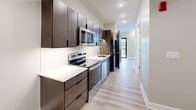 This compact, single-floor unit is thoughtfully designed to maximize functionality within its efficient footprint. The layout, as seen in the floor plan, organizes the space into a galley-style kitchen, a bathroom, and an open living area, all connected by a straightforward corridor flow.

The kitchen is a standout feature, designed in a galley configuration with dark espresso-toned cabinetry extending from floor to ceiling on both sides. White quartz countertops provide a striking contrast against the cabinetry, while a classic white subway tile backsplash adds texture and visual interest. Stainless steel appliances—including an electric range, built-in microwave, dishwasher, and refrigerator—are fully integrated. Light wood-look plank flooring runs throughout the kitchen and into the main living area, lending warmth and continuity.

The living area features neutral gray accent walls, a ceiling fan with integrated lighting, and recessed fixtures that provide even illumination. Natural light enters through a window and a glass-panel exterior door visible from the kitchen corridor.

The bathroom maintains the unit's modern aesthetic with large-format light tile flooring, crisp white walls, a wall-mounted towel bar, and a sleek dark wood vanity. The overall design is clean, contemporary, and efficiently planned throughout.
