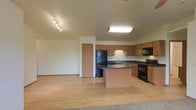 This single-floor residence offers a well-designed open layout that seamlessly connects the kitchen and living areas. The kitchen is a functional centerpiece, equipped with warm honey-toned cabinetry providing ample storage, dark granite countertops, and a central island that serves as both a prep space and casual gathering point. Black appliances, including a refrigerator, range, and built-in microwave, add a sleek contrast to the cabinetry. A flush ceiling light fixture ensures the kitchen is well-lit, while track lighting illuminates the adjacent open area. Light hardwood-style flooring runs throughout the kitchen and connecting spaces, contributing to a clean, cohesive aesthetic.

The living area transitions to plush carpeting, creating a comfortable and distinct zone for relaxation. A ceiling fan with an integrated light fixture provides both air circulation and ambient lighting. Large sliding glass doors flood the room with natural light and open to a private balcony, offering pleasant outdoor views of greenery. The overall ambiance of the space is bright, functional, and inviting, with a neutral palette that suits a variety of décor styles.