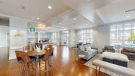 Located on Goodstone Drive in Raleigh, NC, this single-floor community clubroom offers a well-appointed gathering space designed for comfort and socializing. Rich hardwood flooring runs throughout, complementing the soft gray walls, crown molding, and recessed lighting that contribute to the space's polished, contemporary aesthetic.

The open layout flows seamlessly between distinct functional zones. A prominent lounge area anchors one side of the room, featuring a white-mantled fireplace topped with a wall-mounted television, surrounded by stylish gray sofas, accent chairs, and patterned area rugs. Large floor-to-ceiling windows flood the space with natural light and offer direct views of the outdoor pool and surrounding community.

A dedicated café-style bar area is equipped with a commercial-grade coffee machine, granite countertops, warm wood cabinetry, and herringbone tile backsplash—branded as a Starbucks coffee station. Adjacent to this, a bistro-height dining table with walnut chairs provides an ideal casual seating nook beside another window overlooking the pool deck.

Additional seating areas with upholstered benches and accent furniture round out the space, creating a versatile, inviting environment suited for both relaxed lounging and informal gatherings.