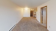This 1-bedroom, 1-bathroom residence is thoughtfully laid out across a single floor, offering a functional and comfortable living environment. The living room is generously proportioned, featuring wall-to-wall carpeting, a baseboard heating system, and a large sliding glass door that fills the space with natural light and provides views of the surrounding outdoor scenery. A modest chandelier adds a touch of warmth overhead. The kitchen is efficiently designed with a galley-style configuration, offering ample white cabinetry for storage, a double stainless steel sink, light-toned countertops, and tile flooring. A range is included, making the kitchen fully equipped for everyday cooking needs. The bedroom continues the carpeted flooring found throughout the living areas and is equipped with a wall-mounted air conditioning unit and a sliding window that frames outdoor views, along with baseboard heating for year-round comfort. The full bathroom features tile flooring, a single-sink vanity with a light stone-finish countertop, a toilet, and a full tub-shower combination with a clean white surround. Warm wood-toned doors and trim accents carry throughout the unit, contributing to a cohesive and inviting aesthetic.