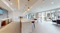 REVL Med Center is a thoughtfully designed, single-floor amenity space that blends contemporary aesthetics with functional social areas. The open-plan layout flows seamlessly from one zone to the next, unified by light tile flooring, crisp white walls, and recessed lighting throughout.

The central gathering area features a striking terrazzo-topped island bar with warm gold pendant lights suspended above, complemented by a full bar backdrop clad in rich blue mosaic tile and warm wood cabinetry. A built-in fireplace anchors one wall, adding an inviting focal point to the space.

Adjacent to the bar, a games area accommodates a ping-pong table and a classic full-length wooden shuffleboard table, with dual wall-mounted screens displaying the REVL Med Center branding. This zone is ideal for casual recreation and social engagement.

A lounge area is defined by a vertical wood slat partition that provides subtle separation while maintaining visual openness. Contemporary seating in bold amber and neutral tones, paired with curated artwork, creates a welcoming, stylish retreat.

Along the perimeter, expansive floor-to-ceiling windows flood the space with natural light, framing outdoor courtyard views. High-top bar tables and seating line the glass wall, offering a bright, airy setting. The overall ambiance is modern, vibrant, and well-appointed for community gathering and recreation.
