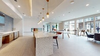 REVL Med Center is a thoughtfully designed, single-floor amenity space that blends contemporary aesthetics with functional social areas. The open-plan layout flows seamlessly from one zone to the next, unified by light tile flooring, crisp white walls, and recessed lighting throughout.

The central gathering area features a striking terrazzo-topped island bar with warm gold pendant lights suspended above, complemented by a full bar backdrop clad in rich blue mosaic tile and warm wood cabinetry. A built-in fireplace anchors one wall, adding an inviting focal point to the space.

Adjacent to the bar, a games area accommodates a ping-pong table and a classic full-length wooden shuffleboard table, with dual wall-mounted screens displaying the REVL Med Center branding. This zone is ideal for casual recreation and social engagement.

A lounge area is defined by a vertical wood slat partition that provides subtle separation while maintaining visual openness. Contemporary seating in bold amber and neutral tones, paired with curated artwork, creates a welcoming, stylish retreat.

Along the perimeter, expansive floor-to-ceiling windows flood the space with natural light, framing outdoor courtyard views. High-top bar tables and seating line the glass wall, offering a bright, airy setting. The overall ambiance is modern, vibrant, and well-appointed for community gathering and recreation.