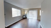 This single-floor space presents a well-appointed interior defined by clean lines and a neutral color palette. The living area is anchored by rich, dark hardwood flooring that extends throughout, providing warmth and visual continuity across the open layout. Walls are finished in a soft gray tone that complements the natural light pouring in through a large picture window, which frames views of surrounding greenery and creates a bright, airy atmosphere.

A full-width sliding glass door fitted with vertical blinds serves as a prominent architectural feature, offering both a secondary source of natural light and direct access to the outdoors. The textured popcorn ceiling adds a classic residential character to the space.

The room is generously proportioned, providing ample space for a variety of furniture arrangements. A wall-mounted heater unit is integrated discreetly into the lower wall, ensuring functional comfort. The overall ambiance is calm and understated, with the interplay of natural light, warm flooring tones, and muted wall colors creating an inviting environment well-suited for everyday living.