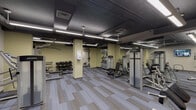 Located at 2009 M E Valentine Dr in Raleigh, NC, this fitness center is a comprehensively equipped single-floor facility designed to support a full range of workout routines. The space features an open, industrial-style aesthetic, with exposed ductwork and dark-painted ceilings contrasted by warm yellow-toned walls and patterned gray carpet tile flooring throughout.

The main workout area is spacious and well-lit by long linear fluorescent fixtures suspended from the ceiling. It is outfitted with a robust selection of Life Fitness strength machines, including cable systems, lat pulldown stations, and bench press setups, as well as a dedicated free weight section with a multi-tier dumbbell rack. A wall-mounted flat-screen TV provides entertainment during workouts, and mirrored panels along one wall enhance the sense of space.

A separate cardio zone houses a row of upright stationary bikes, accessible through frosted-glass double doors. An elliptical machine is also visible in this section. Adjacent to the workout areas is a utility corridor featuring freestanding black changing stalls and access to a storage or utility room, providing practical support amenities. Structural columns are distributed throughout, giving the facility a defined, segmented layout while maintaining a cohesive open feel.