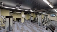 Located at 2009 M E Valentine Dr in Raleigh, NC, this fitness center is a comprehensively equipped single-floor facility designed to support a full range of workout routines. The space features an open, industrial-style aesthetic, with exposed ductwork and dark-painted ceilings contrasted by warm yellow-toned walls and patterned gray carpet tile flooring throughout.

The main workout area is spacious and well-lit by long linear fluorescent fixtures suspended from the ceiling. It is outfitted with a robust selection of Life Fitness strength machines, including cable systems, lat pulldown stations, and bench press setups, as well as a dedicated free weight section with a multi-tier dumbbell rack. A wall-mounted flat-screen TV provides entertainment during workouts, and mirrored panels along one wall enhance the sense of space.

A separate cardio zone houses a row of upright stationary bikes, accessible through frosted-glass double doors. An elliptical machine is also visible in this section. Adjacent to the workout areas is a utility corridor featuring freestanding black changing stalls and access to a storage or utility room, providing practical support amenities. Structural columns are distributed throughout, giving the facility a defined, segmented layout while maintaining a cohesive open feel.