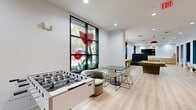 This well-appointed clubhouse is a single-floor amenity space designed to foster community engagement and recreational activity. The open, elongated floor plan is finished with light hardwood-style flooring and bright white walls, creating a clean and energetic backdrop throughout.

The game area features a foosball table and a pool table, positioned near large black-framed windows adorned with vibrant floral artwork that invites natural light and adds a playful artistic touch. Nearby, a lounge area offers a tufted cognac leather sectional sofa, rounded accent chairs, and a ping-pong table, complemented by a wall-mounted flat-screen TV set against a vertical slat accent wall with built-in wood cabinetry beneath.

A centrally located kitchenette serves as the social hub, featuring a wood-and-dark-stone island with mustard yellow barstools, pendant lighting draped with cascading greenery, and a bold mosaic-tiled accent wall. A wall-mounted TV above the counter enhances the space's functionality.

Additional seating zones include a teal and orange booth-style nook with white chairs, ideal for casual dining or conversation. Recessed lighting throughout ensures the space is well-lit, while colorful design accents and curated furnishings give the clubhouse a modern, lively ambiance suited for both relaxation and socializing.