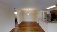 This single-floor residence offers a thoughtfully designed open layout that seamlessly connects the main living and dining areas. The space is anchored by rich, dark hardwood flooring that runs throughout, lending warmth and character to the interior. A textured ceiling adds subtle architectural interest overhead, while neutral wall tones create a clean, versatile backdrop throughout.

The dining area is highlighted by a contemporary pendant light fixture with a geometric frame, providing a focal point that draws the eye upward. A granite-topped counter or bar area is visible along one side, hinting at a well-appointed kitchen nearby. The open passageway leads to an additional room — visible in the background — featuring cabinetry and a window that brings in natural light, suggesting a bathroom or secondary functional space.

The layout benefits from a sense of openness and flow, with wide doorways and clean sight lines connecting each area. Recessed ventilation and standard white trim framing the walls and floors add to the polished, move-in-ready feel. Overall, the space presents a practical yet refined environment, balancing modern fixtures with classic flooring for a timeless residential aesthetic.