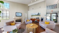 Located in San Diego, CA, this single-floor community clubhouse on Avenida Alcachofa offers a thoughtfully designed gathering space that blends comfort and functionality. The main lounge is the heart of the facility, boasting soaring vaulted ceilings that amplify the sense of openness and airiness throughout. Neutral warm tones on the walls are complemented by plush mid-century modern seating in soft grey, accented with vibrant yellow throw pillows that add a cheerful pop of color. A statement wood-slab coffee table serves as a striking centerpiece, while a classic tile-surround fireplace anchors one end of the room, adding warmth and character. A wall-mounted flat-screen TV provides entertainment, and colorful landscape artwork adorns the walls, enhancing the inviting ambiance. Large French doors and tall windows flood the space with abundant natural light and offer direct views and access to the outdoor areas beyond. A beverage station with a coffee maker adds everyday convenience for residents. Adjacent to the main lounge, a compact and well-equipped business center features dual workstations with desktop computers and ergonomic seating, providing a quiet and focused environment for productivity. Together, these spaces create a versatile and welcoming community hub designed to serve both relaxation and work needs.