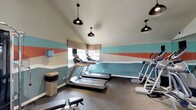 The fitness center at 5427 Lake Murray in La Mesa offers a well-appointed workout space designed for both functionality and visual appeal. The single-floor facility is immediately eye-catching, featuring bold geometric color-block accent walls in teal, coral, and cream tones that lend the space a modern, energetic atmosphere. Vaulted ceilings rise above, creating an open and airy feel, while a series of stylish black pendant lights provide focused overhead illumination throughout the room. Natural light filters in through a window fitted with a woven Roman shade, brightening the space during daytime hours. The flooring consists of durable dark rubber gym matting, ideal for high-traffic fitness use. The facility is equipped with multiple treadmills and elliptical machines, catering to a variety of cardio workout preferences. Additional strength and conditioning equipment is also visible, ensuring a well-rounded fitness experience. A paper towel dispenser and waste receptacle are conveniently positioned for easy access, supporting cleanliness and hygiene. Overall, the space strikes a balance between practical fitness amenities and thoughtful interior design, making it an inviting environment for residents looking to maintain an active lifestyle.