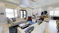 This well-appointed single-floor space exudes a refined yet inviting ambiance, thoughtfully designed to blend comfort with contemporary style. The open lounge area is anchored by generously sized tufted sofas in neutral tones, complemented by richly upholstered accent chairs in bold jewel tones that add a vibrant contrast to the light, airy palette of the room. A tray ceiling with a ceiling fan adds architectural interest overhead, while oversized pendant lighting and recessed fixtures ensure the space is well-lit at all times. Large windows and glass-paneled entry doors flood the interior with natural light, enhancing the sense of openness and connecting the space visually to the outdoors. Light hardwood-style flooring runs throughout, grounded by deep-toned area rugs that define the seating zones. Decorative wall art and sculptural wall accents provide bold focal points, while a dedicated desk area in the background suggests a functional workspace within the same open layout. Every detail — from the layered throw pillows to the curated décor — reflects a deliberate design sensibility that balances elegance with everyday livability.