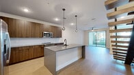 This two-story residence showcases a contemporary design with a cohesive neutral palette and modern finishes throughout. The open-concept main living area seamlessly connects the kitchen and living room, creating a spacious, flowing environment. The kitchen is well-appointed with flat-front walnut-toned cabinetry extending to the ceiling, stainless steel appliances including a French door refrigerator and built-in microwave, and light quartz countertops paired with a clean white tile backsplash. A generous center island with a grey quartz surface and matte black fixtures serves as a functional focal point, illuminated by stylish globe pendant lights overhead. Light wood-look flooring runs throughout the main living spaces, while recessed lighting and a ceiling fan add practical comfort. Large sliding glass doors open to a balcony, drawing in abundant natural light and outdoor views. The bedrooms feature plush carpeting and ceiling fans, with large windows providing additional brightness. A spacious walk-in closet with wrap-around rod systems offers excellent storage capacity. The primary bathroom is finished with large-format light tile flooring, a wood-toned vanity with matte black hardware, and a glass-enclosed walk-in shower with floor-to-ceiling tile surround.