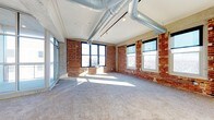 This single-floor space showcases a striking industrial-loft aesthetic that blends raw architectural character with functional design. The interior is defined by prominent exposed brick walls that lend warmth and texture throughout, complemented by polished concrete structural elements and a white ceiling from which large-scale silver ductwork runs openly overhead, reinforcing the industrial character of the space.

The open-plan layout provides a generous, unobstructed floor area covered in neutral gray carpeting, offering a versatile canvas for a variety of uses. Multiple large windows with dark-framed trim are positioned along the brick walls, inviting an impressive amount of natural light that casts dramatic sunlight across the floor throughout the day. A full-height glazed door or window assembly on one side further enhances the brightness and provides an additional connection to the exterior.

Track lighting is installed along the ceiling, offering supplemental illumination to complement the natural light. The overall ambiance is open, airy, and characterful — combining the raw, textured elements of an older building with a clean, updated interior. This space is well-suited for creative, commercial, or professional uses that benefit from a distinctive, light-filled industrial environment.