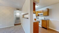 This single-story residence offers a practical and well-organized layout across several distinct areas. The galley-style kitchen is lined on both sides with warm wood-toned cabinetry, dark granite-style countertops, and white appliances including an electric range, dishwasher, and refrigerator. A pass-through window connects the kitchen to the adjacent living area, where a pendant light fixture illuminates the dining space nearby, finished with wood-look flooring. The living room is carpeted and features a ceiling fan with integrated lighting, textured ceilings, and a sliding glass door that opens to a private balcony with outdoor views. A hallway connects the main living areas to the bedroom, which is generously sized with plush carpet, a window overlooking greenery, and access to a walk-in closet outfitted with built-in shelving and hanging rods. The bathroom includes a white tiled tub-surround with shower, a toilet, wood-look flooring, and a vanity with dark countertop. Throughout the unit, neutral wall tones and consistent finishes create a cohesive, functional living environment.