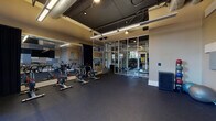 This contemporary fitness facility offers a well-appointed, single-floor layout designed to support a variety of workout styles. The main workout area features durable dark rubber flooring throughout, providing a safe and functional surface for exercise. A row of stationary cycling bikes takes center stage, positioned in front of full-length mirrors that amplify the sense of space and allow users to monitor their form during sessions. The exposed ceiling features industrial-style ductwork and suspended linear lighting fixtures that cast bright, even illumination across the floor.

A striking floor-to-ceiling glass partition wall with a metal frame divides the cycling area from the broader fitness space, creating visual openness while defining distinct training zones. The adjacent area includes modular seating benches, additional exercise equipment, and organized storage cubbies, making it a versatile multi-use space.

Acoustic panels in varying shades of gray and white are mounted on the walls alongside a large flat-screen display, enhancing both the audio environment and visual experience for group classes or individual training sessions. Medicine balls and stability equipment are neatly arranged for easy access. Large windows fitted with blackout curtains allow natural light to filter in while offering the option for a more focused, dimly lit workout environment, with outdoor greenery visible beyond the glass.
