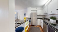 Located at 1317 East St in New Britain, CT, this single-floor residence offers a thoughtfully designed layout with a clean, contemporary aesthetic throughout. The kitchen is equipped with stainless steel appliances — including a refrigerator, dishwasher, and range — complemented by white shaker cabinetry, dark granite countertops, and hardwood flooring. A peninsula with bar seating connects the kitchen to the living area, creating an open, functional flow. The living room is bright and welcoming, featuring windows with blinds that allow natural light to fill the space, a textured ceiling, and room for generous seating arrangements. A secondary sitting area provides additional flexible living space with soft carpeting and neutral tones. The bedrooms are spacious and serene, with carpeted floors, large windows, and a neutral color palette that lends a calm, restful atmosphere. The bathrooms are finished in a clean, minimal style with white vanities, mirrored medicine cabinets, tile accents, and wood-look flooring. Accessibility features such as grab bars are present in the bathrooms. Overall, the residence presents a cohesive modern design with practical finishes and a comfortable, livable ambiance throughout.