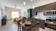 This single-floor residence offers a thoughtfully designed open-concept layout that seamlessly connects the kitchen and living areas into one cohesive, inviting space. The kitchen is a standout feature, showcasing rich dark wood cabinetry that extends to the ceiling, sleek dark granite countertops, and a central island with bar seating — ideal for casual dining or entertaining. Pendant lighting suspended above the island adds warmth and character, while a subway tile backsplash and undermount sink contribute a clean, contemporary aesthetic. The adjoining living area benefits from soaring ceilings and is equipped with a ceiling fan, creating a bright and airy atmosphere. Large windows dressed with patterned curtains allow generous natural light to filter through, enhancing the open feel of the space. Light wood-tone flooring runs throughout, tying the kitchen and living areas together harmoniously. Sliding glass doors lead to a private balcony enclosed by sleek black metal railings, offering views of an open urban landscape under expansive desert skies. The overall ambiance of this residence is modern and well-appointed, balancing functional design with stylish finishes throughout.