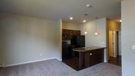 This single-floor residence offers a thoughtfully designed open-concept layout that seamlessly connects the kitchen and living areas. The kitchen is appointed with rich, dark wood cabinetry that extends to the upper walls, providing ample storage while creating a bold visual contrast against the neutral-toned walls. Black appliances, including a refrigerator, range, and microwave, complement the cabinetry for a cohesive, modern aesthetic. A curved kitchen island with a built-in sink and dark countertops serves as the centerpiece of the space, ideal for casual dining or meal preparation. Elegant pendant lights hang above the island, adding warm, focused illumination that enhances the overall ambiance. Recessed lighting in the kitchen further brightens the workspace. The adjoining living area features plush, neutral-toned carpeting that adds warmth and comfort underfoot, offering a welcoming contrast to the dark hardwood-style flooring in the kitchen. Clean lines, neutral wall tones, and a practical layout come together to create a space that is both functional and inviting, suited for everyday living.