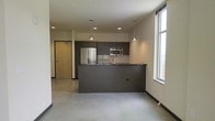 This single-floor space showcases a sleek, modern design centered around an open-concept layout that seamlessly integrates the kitchen and living area. The kitchen is outfitted with dark cabinetry that contrasts sharply against light-toned walls, creating a bold contemporary aesthetic. Stainless steel appliances, including a French door refrigerator and built-in microwave, add a polished, professional quality to the space. A peninsula-style counter extends from the kitchen, offering additional workspace and a natural divider between the cooking and living areas.

Overhead, two pendant lights hang above the peninsula, complementing the recessed lighting throughout to create a layered lighting scheme. The flooring is a smooth, polished concrete that reinforces the industrial-modern character of the space. A large window along one wall floods the interior with natural light, enhancing the open feel of the layout. The entry door, finished in warm wood tones, provides a subtle contrast to the otherwise cool, neutral palette. A tiled backsplash adds texture and detail to the kitchen wall, completing the refined, urban aesthetic.