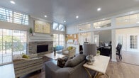 Located at 9010 Markville Dr in Dallas, TX, this single-floor community clubhouse delivers a well-appointed gathering space with a contemporary yet inviting design throughout. The main lounge area serves as the heart of the space, boasting soaring ceilings with clerestory windows that flood the room with natural light. Rich hardwood-style flooring, crown molding, and a striking stone-surround fireplace topped with bold gold-and-white artwork anchor the room with warmth and character. Comfortable gray sofas, patterned accent chairs, and light wood coffee tables create an open, relaxed seating arrangement ideal for socializing. French doors provide direct views and access to an outdoor area. Adjacent to the lounge, a compact kitchenette features granite countertops, a breakfast bar with upholstered stools, gray cabinetry, and black appliances, offering a convenient refreshment space. The business center, finished in a calming slate blue, is equipped with a long granite-topped workstation lined with computers and private dividers, complemented by a round conference table, accent seating, and large windows draped in sheer white curtains. A built-in white display cabinet and coffered ceiling details add architectural interest throughout, making this clubhouse a versatile and well-designed community hub.