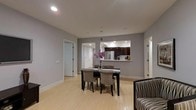 This single-floor space showcases a contemporary open-concept design that seamlessly connects the main living and dining areas. The dining space is anchored by a sleek dark-framed table paired with upholstered grey chairs, complemented by a decorative floral arrangement as a centerpiece. Light hardwood flooring extends throughout, adding warmth against the cool grey wall tones and crisp white baseboards and trim.

A pass-through window opens directly into the kitchen, which features dark cabinetry, stainless steel appliances including a full-sized refrigerator, and a light-colored countertop. Recessed lighting is installed throughout both the main area and kitchen, providing consistent, even illumination across the space.

Multiple doorways are visible, suggesting access to additional rooms beyond the main living area. Clean lines and a neutral color palette define the overall aesthetic, creating a modern and functional environment. A patterned accent chair is partially visible at the edge of the frame, hinting at a connected living area that extends the usable space further.