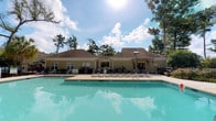 Located at 8439 Dorchester Rd in North Charleston, South Carolina, this single-story community facility serves as a welcoming gathering hub for residents. The centerpiece of the property is a large, pristine outdoor swimming pool with vivid turquoise water, framed by a generous concrete sun deck lined with lounge chairs — ideal for relaxing on warm Carolina days. The clubhouse building features a classic exterior with neutral siding, dark shingled roofing, and a charming pergola-style entryway that adds architectural character to the facade. Multiple windows allow natural light to filter into the interior spaces. The grounds are lushly landscaped, with tall mature trees providing shade and a sense of seclusion while maintaining an open, airy atmosphere. A perimeter fence ensures safety around the pool area, while additional recreational elements are visible along the poolside. The overall ambiance is inviting and resort-like, blending functional amenity spaces with comfortable outdoor living in a well-maintained, community-oriented setting.