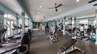 Located at 8230 Stonebrook Terr in Raleigh, NC, this single-floor fitness center offers a well-rounded workout environment designed to accommodate a wide range of exercise needs. The space is finished with durable rubber flooring throughout and features a distinctive color palette of soft blue and teal accent walls paired with crisp white trim and columns, creating a clean and energizing atmosphere.

The main workout area is generously proportioned, housing an extensive array of strength training machines, free weights, and functional training equipment including TRX suspension systems. Large arched windows along one wall flood the cardio section with abundant natural light and provide views of the outdoor surroundings, while a row of treadmills and ellipticals is positioned to take full advantage of this feature. Ceiling fans are installed throughout to maintain comfortable airflow during workouts.

Full-length mirrors line portions of the walls, enhancing the sense of openness and allowing users to monitor their form. A dedicated open area near the entrance supports functional and bodyweight training. Additional amenities include a wall-mounted television, a water fountain station, and access to an adjacent room that appears to serve as a supplementary activity or multipurpose space. The overall layout is thoughtfully organized to ensure ample room between equipment stations.