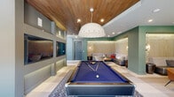 Located at 3704 Governors Dr SW in Huntsville, Alabama, this single-story community clubhouse delivers an impressive blend of modern design and social functionality. The centerpiece is a soaring double-height lobby distinguished by dramatic circular and cascading pendant lighting, large-format tile flooring in a checkered pattern, and floor-to-ceiling windows that flood the space with natural light. A bold geometric mosaic feature wall anchors the main lounge area, which is furnished with contemporary sofas and accent chairs arranged around stylish coffee tables.

Adjacent to the main lounge is a dedicated wet bar area with dark cabinetry, a built-in sink, under-counter appliances, and a richly veined stone countertop — ideal for entertaining. A large-format TV is mounted within the geometric tile surround, adding a media element to the social hub.

The game lounge is a standout feature, showcasing a full-size pool table beneath a warm wood-paneled ceiling and an oversized sculptural pendant light. Twin flat-screen TVs are recessed into the wall, and a shuffleboard table runs along the window line. Plush banquette seating lines the lounge perimeter, complemented by textured wall panels, exposed brick accents, and ambient LED lighting that create an inviting, upscale atmosphere throughout.
