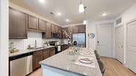 This one-bedroom, one-bathroom unit is thoughtfully designed to maximize functionality and style across a single floor. The open-concept kitchen and living area serves as the heart of the home, where a large granite-topped island with bar seating takes center stage. Rich, medium-toned cabinetry with a classic subway tile backsplash lines the kitchen walls, complemented by a full suite of stainless steel appliances, including a side-by-side refrigerator, electric range, and built-in microwave. Light wood-style flooring runs throughout the main living area, enhancing the bright, airy atmosphere. The living room receives natural light through a glass door and large windows, fitted with elegant floor-length curtains, while a ceiling fan adds practicality. The bedroom is carpeted and features generous window coverage, providing soft natural light. An adjacent walk-in closet offers wire shelving and ample hanging space. The full bathroom connects directly to the bedroom and includes a granite-topped vanity with dark cabinetry, wood-style flooring, and a tub-shower combination with clean white tile surround, completing this well-finished residential unit.