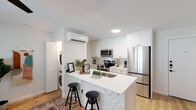 This well-designed single-floor unit offers a thoughtful layout that maximizes functionality and style throughout. The heart of the home is a contemporary open-concept kitchen outfitted with sleek white cabinetry extending to the ceiling, stainless steel appliances including a refrigerator, range, and built-in microwave, and a striking peninsula island featuring a bold geometric tile pattern on its facade and a clean white countertop with bar seating. Light wood flooring runs continuously throughout, lending warmth and visual cohesion to the space. A wall-mounted mini-split system ensures year-round climate comfort. Tucked conveniently behind a louvered door adjacent to the kitchen is a stacked washer and dryer, keeping laundry functions neatly integrated. The living area accommodates comfortable seating and benefits from a large window fitted with wood blinds that allow for natural light control, complemented by a ceiling fan overhead. The bedroom is a serene retreat, featuring its own mini-split unit, a ceiling fan, and a window with blinds. It connects directly to a private bathroom appointed with bold black-and-white geometric tile in the tub surround, a vanity with white countertop, and coordinating fixtures. The overall aesthetic is clean, modern, and cohesive throughout.