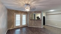 This single-story space offers an open and functional layout highlighted by rich, dark wood-style flooring that runs throughout the main living area. The living room serves as the central gathering space, featuring a decorative ceiling light fixture that casts a warm, ambient glow across the textured ceiling. A set of French doors with white horizontal blinds allows natural light to filter into the room while providing access to the exterior.

A standout architectural detail is the pass-through window connecting the living area to the kitchen, complete with a dark-toned bar counter that doubles as a convenient serving or casual dining surface. This opening frames a view into the kitchen, where white cabinetry, a decorative tile backsplash, and a black refrigerator are visible, offering a sense of continuity between the two spaces.

The neutral gray-toned walls provide a versatile backdrop throughout, complementing the warm flooring tones. A hallway visible from the main living area suggests additional rooms beyond, contributing to the overall sense of a well-connected, efficiently designed single-floor layout. The space balances practicality with accessible design details.