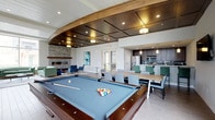 This single-floor community clubroom is thoughtfully designed to serve as a premier social and recreational hub. The space is anchored by a full-sized billiards table set beneath a striking curved wooden archway that frames a recessed coffered ceiling with warm pendant lighting, giving the area a polished, upscale feel. Adjacent to the game area, a spacious dining section features long communal tables with ample seating, perfect for group gatherings or events. A fully equipped kitchen runs along the back wall, complete with stainless steel appliances, dark cabinetry, and pendant lighting, offering a functional space for catering or casual cooking. To one side, a comfortable lounge area provides a more relaxed setting, centered around a modern stone-surround fireplace with built-in shelving and flanked by plush seating in rich blue tones. Natural light filters in through exterior-facing windows and a glass door, brightening the crisp white walls and light tile flooring throughout. The overall ambiance balances contemporary design with welcoming warmth, making this clubroom a versatile and inviting destination for recreation, dining, and social connection.