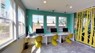 This single-floor community space is thoughtfully designed to support both productivity and collaboration, boasting a lively, contemporary aesthetic throughout. The computer lounge is a standout area, featuring a long white built-in desk lined with iMac workstations, complemented by bold chartreuse Panton-style chairs and vivid teal accent walls. Three large windows flood the room with natural light, while sleek gold pendant lights hang overhead, adding a modern touch. A striking yellow laser-cut room divider — shaped like a grove of trees — serves as a dramatic architectural partition between the computer area and the adjacent spaces, doubling as a work of art. The conference room is defined by a frosted glass sliding wall, offering privacy while maintaining visual connectivity to the surrounding lounge. Inside, a white rectangular table is surrounded by warm wood-toned chairs, and a wall-mounted display screen is set into a rich wood-paneled feature wall. Colorful framed artwork adds personality to the space. Patterned, multi-toned carpeting runs throughout, tying the various zones together. Large windows in the conference area frame outdoor greenery, enhancing the bright and welcoming atmosphere. Overall, the space strikes a balance between functional design and creative expression.
