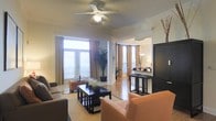 This single-story space showcases an open-concept design that connects the living and dining areas in a cohesive, flowing layout. The living area is anchored by warm caramel-toned seating arranged around a dark wood coffee table, creating an inviting and functional gathering space. Crown molding lines the ceiling throughout, adding a refined architectural detail, while a ceiling fan with a central light fixture provides both airflow and ambient lighting.

Large windows dressed with warm golden curtains allow generous natural light to fill the space, enhancing the bright, airy atmosphere. A tall indoor plant adds a natural, organic element to the corner of the room. The flooring transitions from a neutral area rug in the living space to light-toned hard flooring that extends into the dining area.

The adjacent dining area features a bar-height table paired with dark, sleek bar stools, illuminated by an elegant chandelier overhead. A large dark armoire or entertainment unit stands prominently along one wall, offering substantial storage. The overall ambiance is warm, polished, and welcoming, combining contemporary furnishings with classic architectural touches.