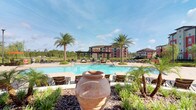 The Pool 360 area is a thoughtfully designed outdoor amenity space that combines resort-style recreation with refined architectural details. At its center sits a generously sized swimming pool with clear blue water, framed by a wide pool deck lined with chaise lounges and surrounded by swaying palm trees and tropical plantings that reinforce a warm, leisurely atmosphere.

A prominent pergola structure anchors the space, featuring bold dark-toned beams supported by classic white columns. Ceiling fans mounted beneath the pergola ensure comfortable airflow, making the shaded areas functional in warm weather. The pergola connects to a clubhouse-style building clad in natural stone, with large windows that reflect the pool and surrounding greenery. Adjacent covered seating areas are furnished with wicker sofas adorned with vibrant orange cushions and accent pillows, creating inviting conversation nooks.

Underfoot, patterned paver stonework extends throughout the patio and walkways, adding texture and visual interest. Ornamental planters, including a large terra cotta urn, serve as focal points within the landscaping beds. The surrounding multi-story residential buildings are visible in the backdrop, grounding the amenity within its community context while open skies above contribute to an airy, expansive ambiance.