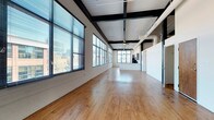 This single-floor space showcases a compelling industrial aesthetic through its open-plan layout and architectural detailing. The expansive main area is anchored by warm, natural hardwood floors that run the length of the space, reflecting light and adding a sense of warmth to the industrial surroundings.

Overhead, exposed black steel beams contrast sharply against a white-painted ceiling, lending the space a raw, loft-like character. A ceiling fan is centrally mounted, contributing to air circulation throughout the generous open area. Visible ductwork and utility elements reinforce the industrial aesthetic.

A continuous row of large, multi-pane windows lines one wall, fitted with horizontal blinds and flooding the interior with natural light. These windows frame elevated views of the surrounding urban landscape, including glimpses of city buildings and mature trees.

Toward the rear of the space, a partial partition wall defines a secondary area, suggesting a kitchen or utility zone, subtly separated from the main open floor. A door on the far wall provides additional access.

Overall, the space conveys a versatile, open ambiance well-suited for creative, commercial, or collaborative use.