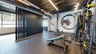 Located at 18807 Eighth Ave NE in Shoreline, Washington, this fitness center offers a spacious, single-level layout designed to accommodate a comprehensive range of workout activities. The space features an industrial-modern aesthetic, with exposed ductwork and structural piping running across the ceiling, complemented by sleek linear LED lighting that illuminates the floor throughout.

The main strength training area is equipped with an extensive array of cable machines, multi-functional racks, resistance stations, adjustable benches, and a full dumbbell rack, providing ample options for both beginners and advanced athletes. A dedicated cardio zone features rows of elliptical trainers positioned near large windows that invite natural light and views of the surrounding greenery, creating an open and energizing atmosphere.

A separate cycling and rowing area showcases stationary bikes and rowing machines alongside wall-mounted flat-screen TVs, while a bold mountainscape mural adds a dynamic visual anchor to the space. Additional zones include a functional training area with TRX suspension equipment, resistance bands, and medicine balls, as well as a quieter alcove accented by decorative wall art. Large-format tile flooring throughout provides both durability and a polished, cohesive appearance. Built-in shelving units offer convenient storage for personal belongings.