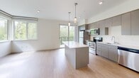 Located at 18475 Redmond Way in Redmond, WA, this single-story residence offers three bedrooms, two bathrooms, and a thoughtfully designed layout. The open-concept kitchen and living area serves as the heart of the home, featuring a sleek center island with a white quartz countertop, modern gray cabinetry, stainless steel appliances including a range, microwave, and refrigerator, and pendant lighting overhead. Light wood-style flooring runs throughout the main living space, and a sliding glass door leads to a private balcony overlooking lush, mature greenery. The bedrooms are carpeted and bathed in natural light through large windows that frame tranquil tree-canopy views — a hallmark of the Pacific Northwest setting. The primary bedroom offers a comfortable footprint with ample wall space. Both bathrooms are finished in a clean, contemporary style; one features a gray vanity with a white countertop, a large mirror, and wood-look flooring, while the second includes a full tub-shower combination. Crisp white walls and recessed lighting throughout contribute to a bright, neutral ambiance that complements the natural surroundings of this Redmond location.