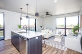 This contemporary single-floor unit showcases a thoughtfully designed open-concept layout that seamlessly connects the kitchen, dining, and living areas. The kitchen is a standout feature, centered around a large island with a white quartz countertop, an undermount sink, and a two-tone design pairing navy-blue lower cabinetry with crisp white upper cabinets. Stainless steel appliances, pendant lighting, and an exposed ductwork detail lend the space an industrial-modern character. Light hardwood flooring flows throughout, enhancing the bright, airy atmosphere. The living area benefits from floor-to-ceiling windows that frame sweeping urban views, flooding the space with natural light. A private balcony extends the living space outdoors. The unit includes multiple bedrooms, each featuring ceiling fans, recessed lighting, and hardwood flooring. One bedroom offers a large window with outdoor views, while another presents a clean, versatile layout. The two bathrooms are finished with warm wood-tone flooring, white surrounds, and dark matte fixtures, offering a clean and cohesive aesthetic. An additional flexible room with recessed lighting and a ceiling fan provides space adaptable to various needs. Overall, the unit delivers a polished, modern ambiance with functional design throughout.