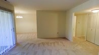 This single-floor residence presents a straightforward and functional layout designed for comfortable everyday living. The main living area is generously proportioned, featuring wall-to-wall carpeting in a warm, neutral beige tone that extends throughout the space, providing a soft and cohesive foundation underfoot. The walls are finished in a light, creamy off-white palette, contributing to a bright and airy atmosphere throughout.

The living area is defined by a partial interior wall that creates a subtle sense of separation while maintaining an open-concept feel between spaces. Smooth, flat ceilings run throughout, and the overall interior is clean and unfurnished, making it easy to envision a variety of furniture arrangements and personal styles.

A hallway visible from the main living space leads to additional rooms, suggesting a well-connected floor plan with accessible transitions between areas. The neutral finishes — from the carpet to the painted walls — provide a versatile backdrop, well-suited to a range of interior design preferences. The space presents a blank canvas that is ready for personalization, with practical, easy-to-maintain finishes throughout. A pendant light fixture is visible in one corner, hinting at thoughtful lighting placement across the home.