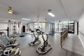 Located at 131 Ponce de Leon Ave NE in Atlanta, GA, this fitness center offers a generously sized, single-floor exercise space designed to accommodate a wide range of workout styles. The layout is open and thoughtfully organized, with durable rubber flooring throughout that provides both safety and comfort underfoot.

Natural light pours in through large, floor-to-ceiling black-framed windows that line the perimeter, offering views of the surrounding Atlanta streetscape and creating an energizing atmosphere. Ceiling fans and recessed lighting complement the brightness, ensuring the space is well-lit at all hours.

The exercise room is comprehensively equipped with multiple Peloton-style cycling bikes, a cable and functional training rig, an elliptical machine, and a flat-screen monitor for guided workouts. A dedicated free weights area features a full rack of colorful kettlebells in varying weights alongside medicine balls and stability equipment, catering to strength and conditioning training. Stretching and group fitness accessories, including exercise mats and stability balls, are also available.

A sleek metal railing system defines the entry staircase area, adding an architectural detail that connects the fitness space to the rest of the building. The clean white walls and neutral color palette create a modern, professional environment that is both functional and visually appealing.