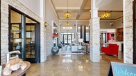 Located at 915 Robert Rose Dr in Murfreesboro, Tennessee, this sophisticated single-floor clubhouse lounge delivers an upscale communal experience through thoughtful architectural design and bold interior styling. The expansive open-plan space is anchored by soaring coffered and tray ceilings that amplify the sense of volume, while gleaming polished concrete floors reflect abundant natural light streaming in through large windows and glass entry doors. Stately stacked stone columns punctuate the floor plan, lending a refined yet organic texture that contrasts elegantly with the crisp white walls and trim. A central lounge area features plush red seating, neutral wingback chairs, and patterned area rugs, creating dynamic gathering zones with distinct character. A boldly accented media nook with a vibrant red accent wall, wall-mounted television, and modular ottomans offers a comfortable retreat for entertainment. The space also includes a sleek counter bar area with bar seating, a reclaimed wood feature wall with built-in monitors, and pendant lighting that adds warmth and intimacy. A shuffleboard table provides a playful recreational focal point, while curated artwork and decorative wall pieces contribute to the space's contemporary aesthetic. Warm pendant and drum light fixtures throughout complete the inviting, community-centered ambiance.
