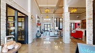 Located at 915 Robert Rose Dr in Murfreesboro, Tennessee, this sophisticated single-floor clubhouse lounge delivers an upscale communal experience through thoughtful architectural design and bold interior styling. The expansive open-plan space is anchored by soaring coffered and tray ceilings that amplify the sense of volume, while gleaming polished concrete floors reflect abundant natural light streaming in through large windows and glass entry doors. Stately stacked stone columns punctuate the floor plan, lending a refined yet organic texture that contrasts elegantly with the crisp white walls and trim. A central lounge area features plush red seating, neutral wingback chairs, and patterned area rugs, creating dynamic gathering zones with distinct character. A boldly accented media nook with a vibrant red accent wall, wall-mounted television, and modular ottomans offers a comfortable retreat for entertainment. The space also includes a sleek counter bar area with bar seating, a reclaimed wood feature wall with built-in monitors, and pendant lighting that adds warmth and intimacy. A shuffleboard table provides a playful recreational focal point, while curated artwork and decorative wall pieces contribute to the space's contemporary aesthetic. Warm pendant and drum light fixtures throughout complete the inviting, community-centered ambiance.