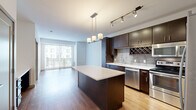 Located at 5245 Wayzata Blvd in St. Louis Park, MN, this well-appointed one-bedroom, one-bathroom residence offers a thoughtfully designed open-concept layout. The combined kitchen and living area serves as the heart of the home, showcasing rich dark-espresso cabinetry, a tiled backsplash with a built-in wine rack, stainless steel appliances including a refrigerator, range, microwave, and dishwasher, and a center island with a white quartz countertop and elegant pendant lighting overhead. Track lighting above the kitchen adds functional brightness, while warm hardwood-style flooring flows throughout the main living areas. The living room is bathed in natural light from large floor-to-ceiling windows and sliding glass doors that open to a balcony with exterior views. The bedroom features plush carpeting, a generous window with outdoor views, and a dedicated closet for storage. The bathroom is finished with a white-tiled tub and shower surround, a dark wood vanity with a white countertop, and hardwood-style flooring that ties the space together. A built-in desk area near the kitchen provides a convenient workspace. Clean white walls and modern finishes throughout lend the space a crisp, contemporary ambiance.