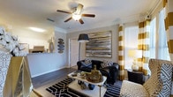 This single-floor residence combines contemporary style with comfortable living across a well-appointed open-concept layout. The living area serves as the centerpiece of the home, featuring rich wood-style flooring that grounds the space with warmth and durability. A ceiling fan with integrated lighting anchors the room from above, providing both function and a finishing architectural touch to the smooth, light-toned ceiling. Large windows dressed with bold gold and white striped curtains invite natural light into the space, enhancing the bright and airy atmosphere. The living room is thoughtfully designed with a striking large-scale abstract wall painting in gold, black, and gray tones, complementing the dark upholstered seating and patterned area rug beneath. A sleek coffee table with gold-tone accents adds a modern, curated feel to the arrangement. Crown molding along the upper walls adds a refined architectural detail throughout. Adjacent to the living area, the kitchen is visible over a breakfast bar-style counter, featuring white cabinetry and under-cabinet lighting, creating a seamless flow between cooking and living spaces. The overall ambiance is bold yet sophisticated, blending geometric patterns, metallic accents, and carefully chosen furnishings into a cohesive and inviting interior.