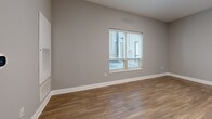 Located at 255 S High St in the heart of downtown Columbus, Ohio, this efficiently designed single-floor unit offers a clean, modern aesthetic with thoughtful finishes throughout. The open living area is anchored by warm hardwood-style flooring, neutral gray walls, and a window fitted with blinds that allows natural light to filter in while offering views of the surrounding urban environment. The kitchen is well-appointed with crisp white shaker-style cabinetry, gray quartz-look countertops, stainless steel appliances including a range, over-the-range microwave, and dishwasher, and a geometric hexagonal tile backsplash that adds a contemporary design touch. A dedicated dining or flex area adjacent to the kitchen provides additional functional space. The bathroom features a white vanity with gold-toned hardware, an undermount sink, neutral stone-look countertop, and a large mirror, all finished in a clean, modern palette consistent with the rest of the unit. The hallway connects the spaces efficiently, with a white paneled entry door visible from the bathroom. Hardwood-style flooring flows consistently throughout, tying the spaces together with a cohesive and polished look ideal for urban living.