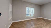 Located at 255 S High St in the heart of downtown Columbus, Ohio, this efficiently designed single-floor unit offers a clean, modern aesthetic with thoughtful finishes throughout. The open living area is anchored by warm hardwood-style flooring, neutral gray walls, and a window fitted with blinds that allows natural light to filter in while offering views of the surrounding urban environment. The kitchen is well-appointed with crisp white shaker-style cabinetry, gray quartz-look countertops, stainless steel appliances including a range, over-the-range microwave, and dishwasher, and a geometric hexagonal tile backsplash that adds a contemporary design touch. A dedicated dining or flex area adjacent to the kitchen provides additional functional space. The bathroom features a white vanity with gold-toned hardware, an undermount sink, neutral stone-look countertop, and a large mirror, all finished in a clean, modern palette consistent with the rest of the unit. The hallway connects the spaces efficiently, with a white paneled entry door visible from the bathroom. Hardwood-style flooring flows consistently throughout, tying the spaces together with a cohesive and polished look ideal for urban living.