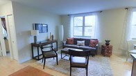 This single-floor space offers a well-designed and inviting living environment with a clean, contemporary aesthetic. The living area serves as the centerpiece of the space, featuring light sage-green walls that create a calm, neutral backdrop for the modern furnishings. A large window dressed with sheer white curtains floods the room with natural light and provides views of the surrounding urban environment. The flooring is light hardwood, adding warmth and continuity throughout the space. The living area is anchored by a plush, light-colored area rug layered over the hardwood, upon which a charcoal-gray sofa adorned with rust and cream accent pillows is tastefully arranged. A dark wood console table hosts a wall-mounted flat-screen television and decorative accents, while a pair of dark wood dining chairs and a coffee table complete the seating arrangement. A slender floor lamp adds a sculptural element alongside the sofa. The overall ambiance is modern yet comfortable, with a cohesive color palette of warm neutrals and earthy tones that contribute to a welcoming atmosphere throughout the space.