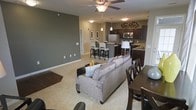 This single-floor residence showcases an open-concept layout that seamlessly integrates the living, dining, and kitchen areas into one cohesive space. The living area is anchored by a plush, light gray sectional sofa accented with decorative pillows, complemented by a ceiling fan with a built-in light fixture overhead. Neutral carpet flooring runs throughout the living and dining zones, while the walls are finished in a muted sage green tone that lends a calm, contemporary feel to the interior.

The dining area is positioned adjacent to the living space, furnished with a dark wood table and matching chairs, creating a natural transition between the two zones. The kitchen is defined by dark espresso cabinetry, granite-look countertops, and stainless steel appliances including a refrigerator and microwave. A kitchen island with bar-height seating and pendant lighting above provides both additional prep space and casual dining options. Decorative wall art adds personality above the upper cabinetry. The overall ambiance is modern and well-appointed, with a cohesive color palette and practical, open flow throughout.